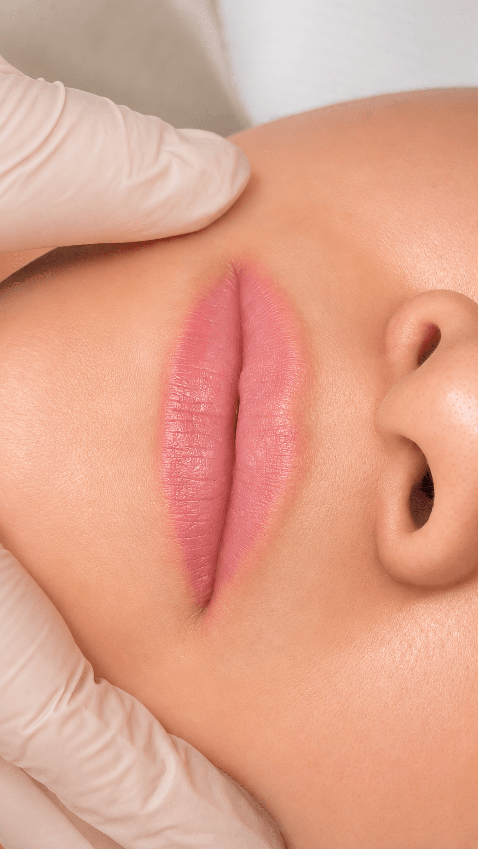 Labio con color aplicado, tono rosado suave, con la mano enguantada de la profesional apoyada sobre la frente.